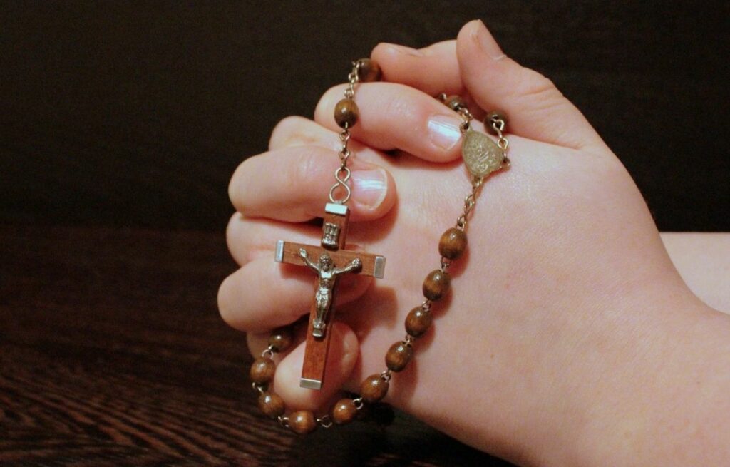 Imagem mostra mãos em oração com terço - oração da manhã baseada no pai nosso